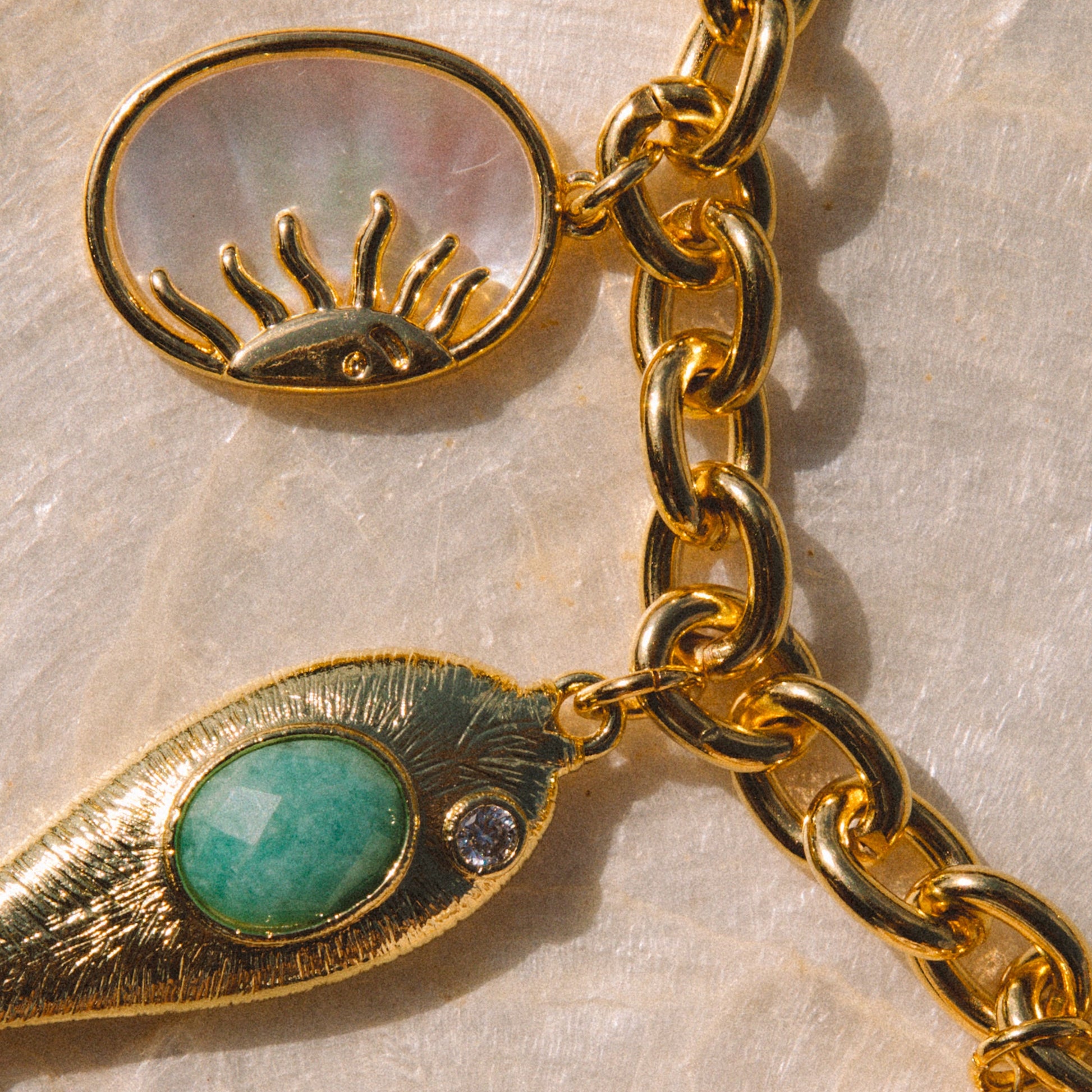 Gold-tone charm bracelet featuring a seashell charm, a textured gold leaf charm with a green oval stone, a mother-of-pearl sun charm, and a gold chain on a textured beige background.