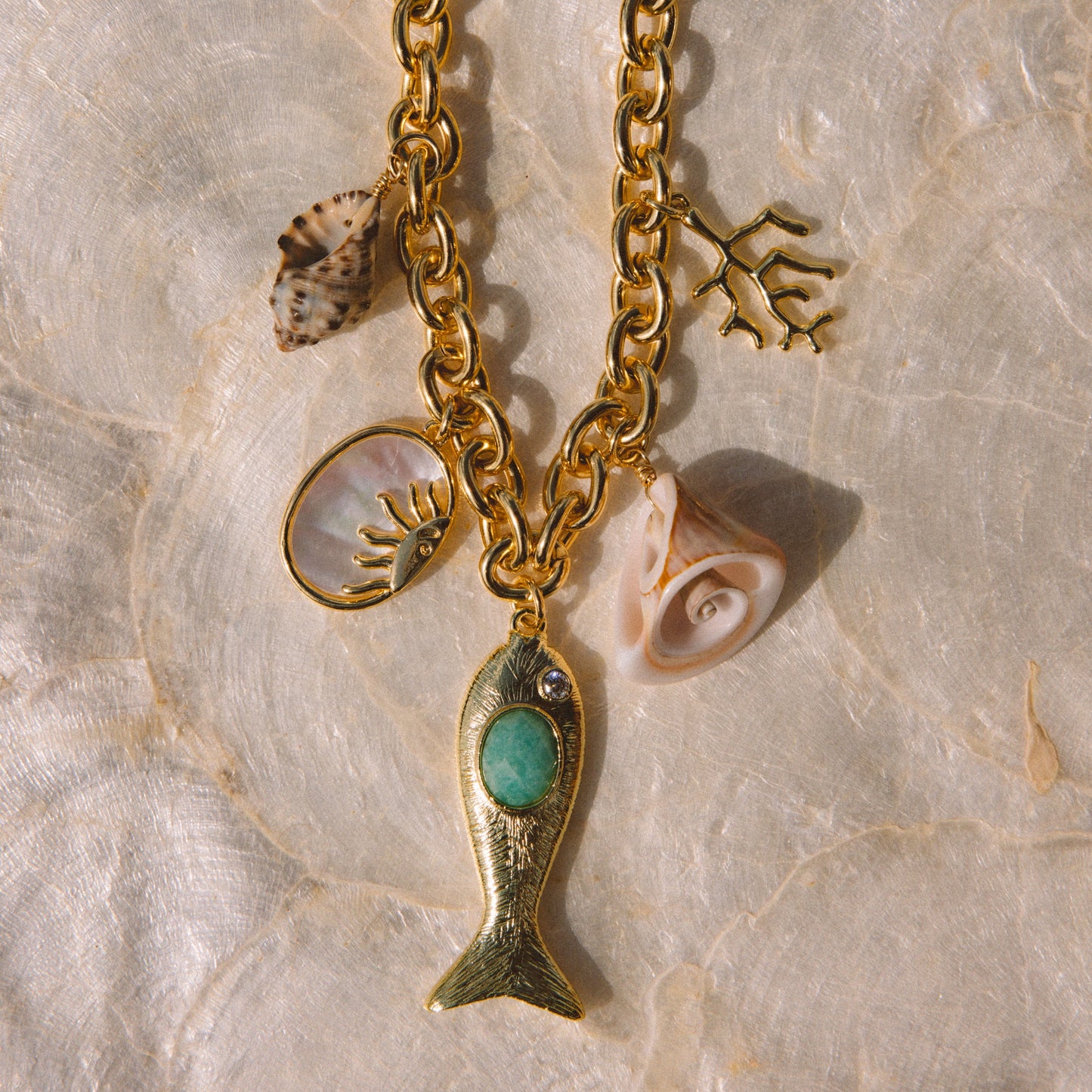 Gold-tone charm necklace featuring a fish pendant with a turquoise center, seashell charms, and coral accents, displayed on a textured beige stone surface.