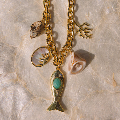 Gold-tone charm necklace featuring a fish pendant with a turquoise center, seashell charms, and coral accents, displayed on a textured beige stone surface.