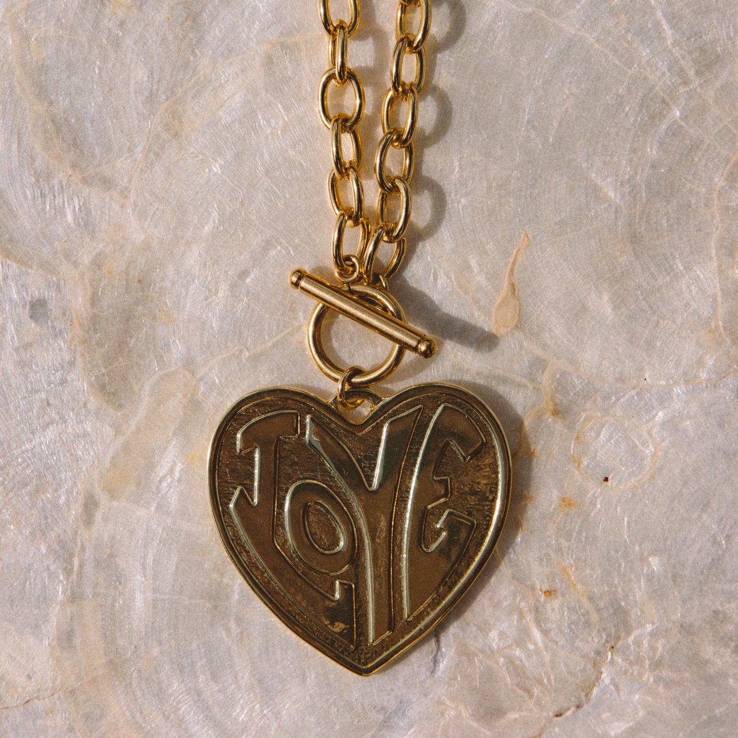 A gold heart-shaped pendant necklace with an engraved design and a toggle clasp, displayed on a textured white stone surface.