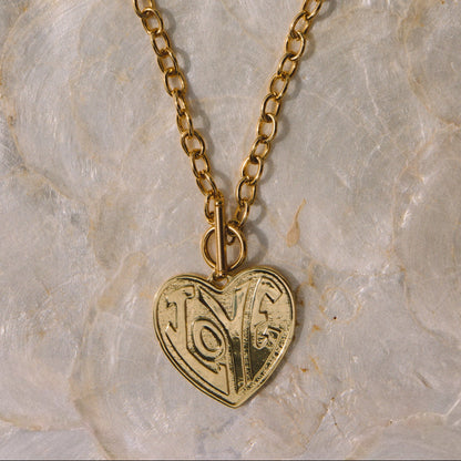 A gold-toned double-strand chain necklace with a heart-shaped pendant featuring engraved swirl patterns, displayed on a textured white stone surface.