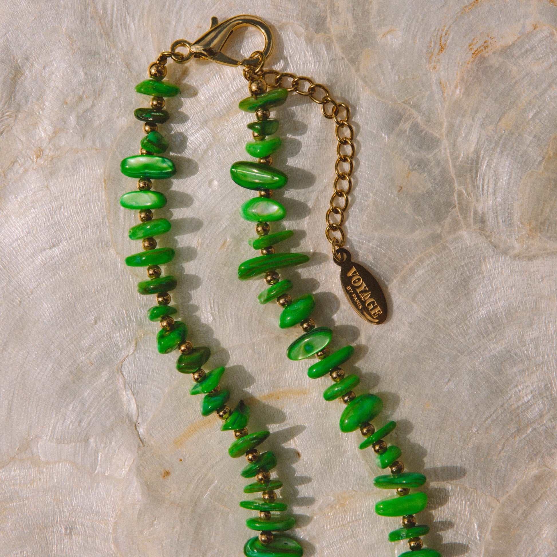A beaded necklace with vibrant green stone chips and small gold spacer beads, featuring a gold-tone lobster clasp, extension chain, and a small oval charm engraved with 'VOYAGE BY PARIS'. The necklace is laid flat on a textured, light beige stone surface.