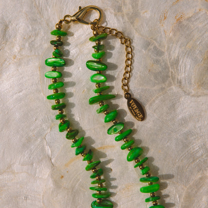 A beaded necklace with vibrant green stone chips and small gold spacer beads, featuring a gold-tone lobster clasp, extension chain, and a small oval charm engraved with 'VOYAGE BY PARIS'. The necklace is laid flat on a textured, light beige stone surface.