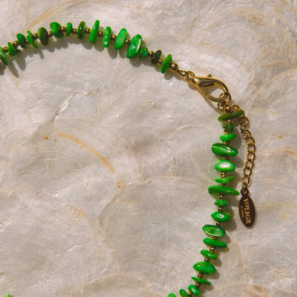 A green beaded necklace with small, irregular disc-shaped beads and a gold-tone clasp, displayed on a textured beige stone surface.