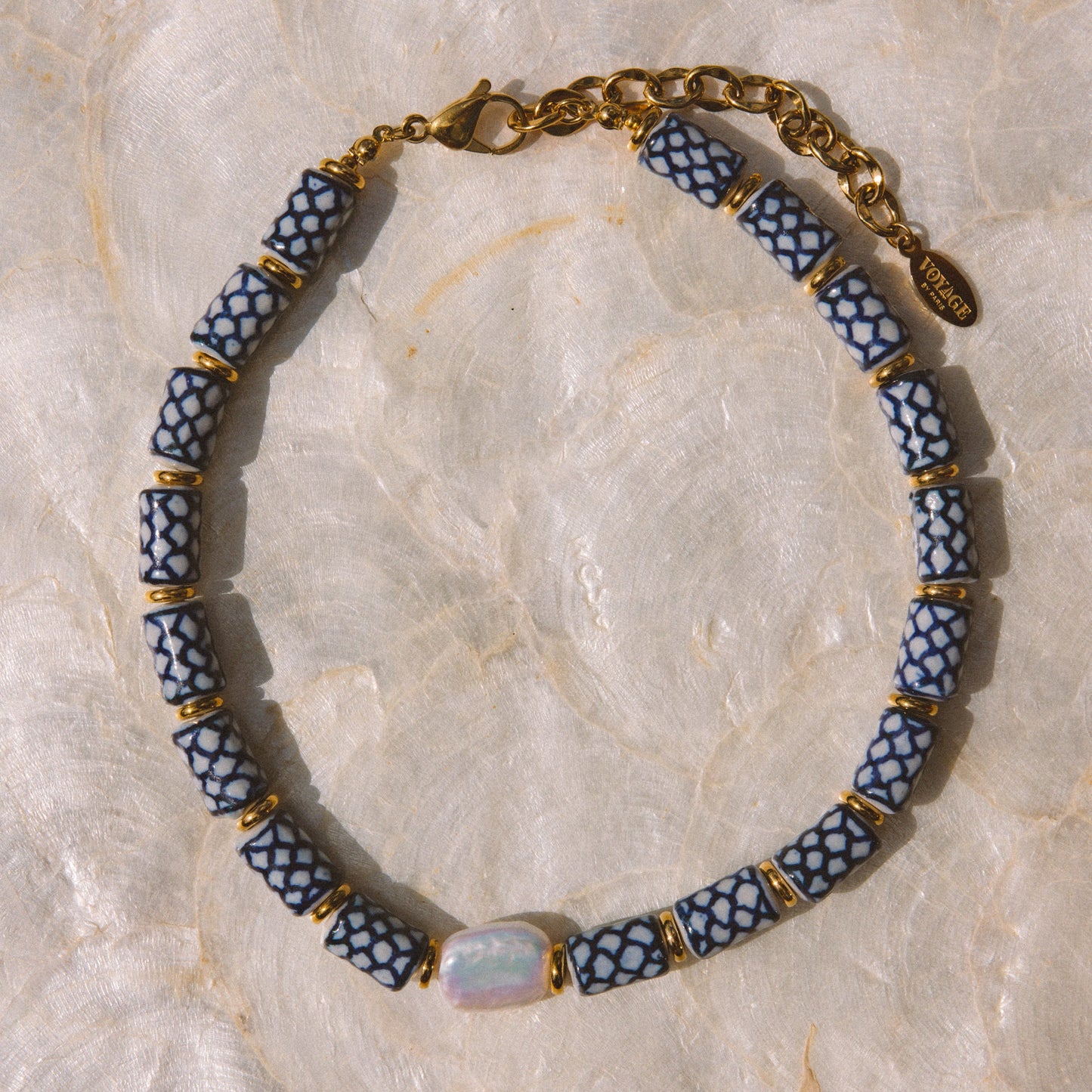A beaded necklace with alternating blue and white patterned cylindrical beads, separated by small gold spacer rings, with a gold chain clasp and a central iridescent oval stone, displayed on a light beige marbled surface.