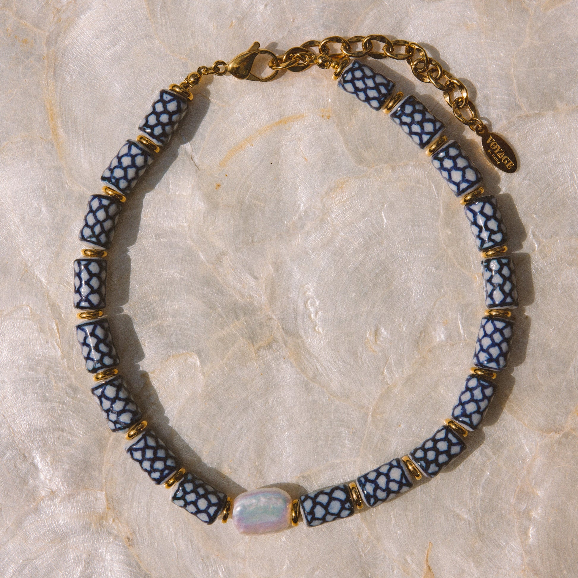 A beaded necklace with alternating blue and white patterned cylindrical beads, separated by small gold spacer rings, with a gold chain clasp and a central iridescent oval stone, displayed on a light beige marbled surface.