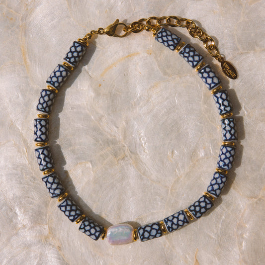 A beaded necklace with alternating blue and white patterned cylindrical beads, separated by small gold spacer rings, with a gold chain clasp and a central iridescent oval stone, displayed on a light beige marbled surface.