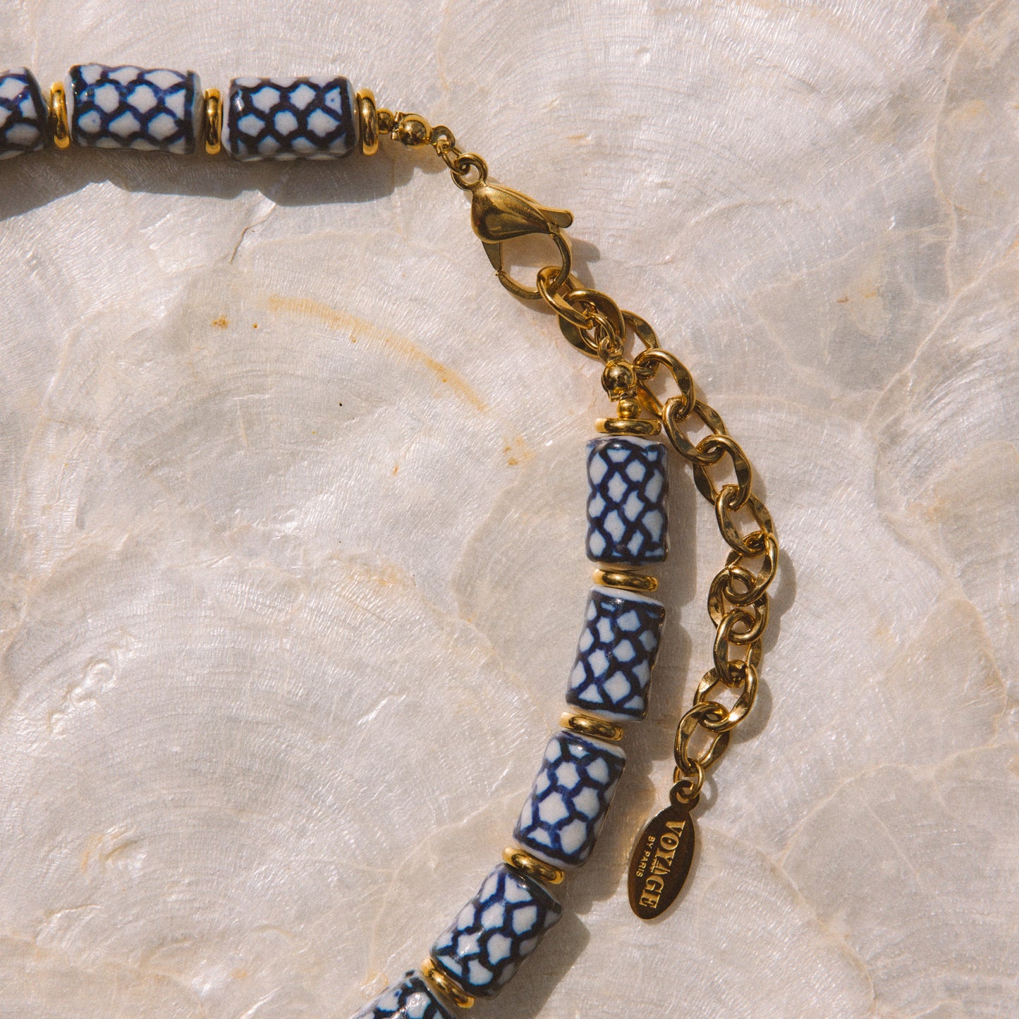 A close-up of a beaded necklace with cylindrical blue and white patterned beads separated by gold spacer rings, connected by a gold chain and clasp, laid on a light marbled surface.