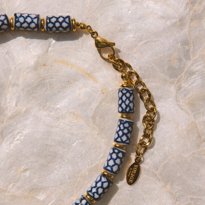 A close-up of a beaded necklace with cylindrical blue and white patterned beads separated by gold spacer rings, connected by a gold chain and clasp, laid on a light marbled surface.