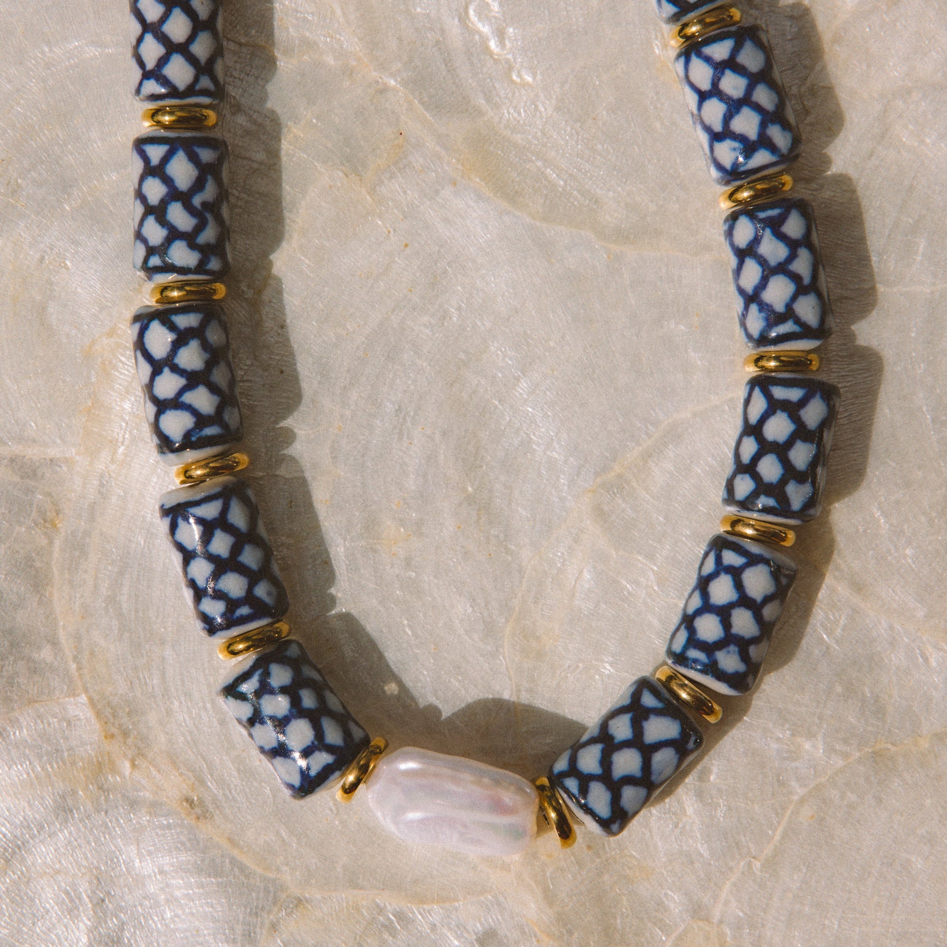 A beaded necklace with blue and white patterned cylindrical beads separated by small gold spacer rings, laid on a light beige textured surface. A white shell is threaded through the center bead.