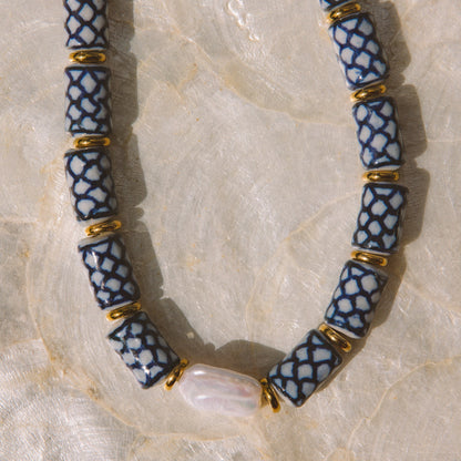 A beaded necklace with blue and white patterned cylindrical beads separated by small gold spacer rings, laid on a light beige textured surface. A white shell is threaded through the center bead.