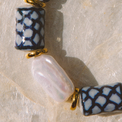 A close-up of a beaded strand featuring dark blue and white patterned cylindrical beads, a smooth white oval bead, and small gold spacer rings, laid on a light stone surface.