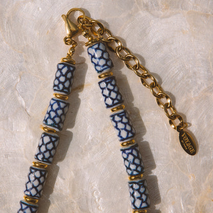 A close-up of a beaded jewelry chain featuring blue and white patterned ceramic beads separated by gold spacer rings, with a gold lobster clasp and extension chain on a textured light beige surface.