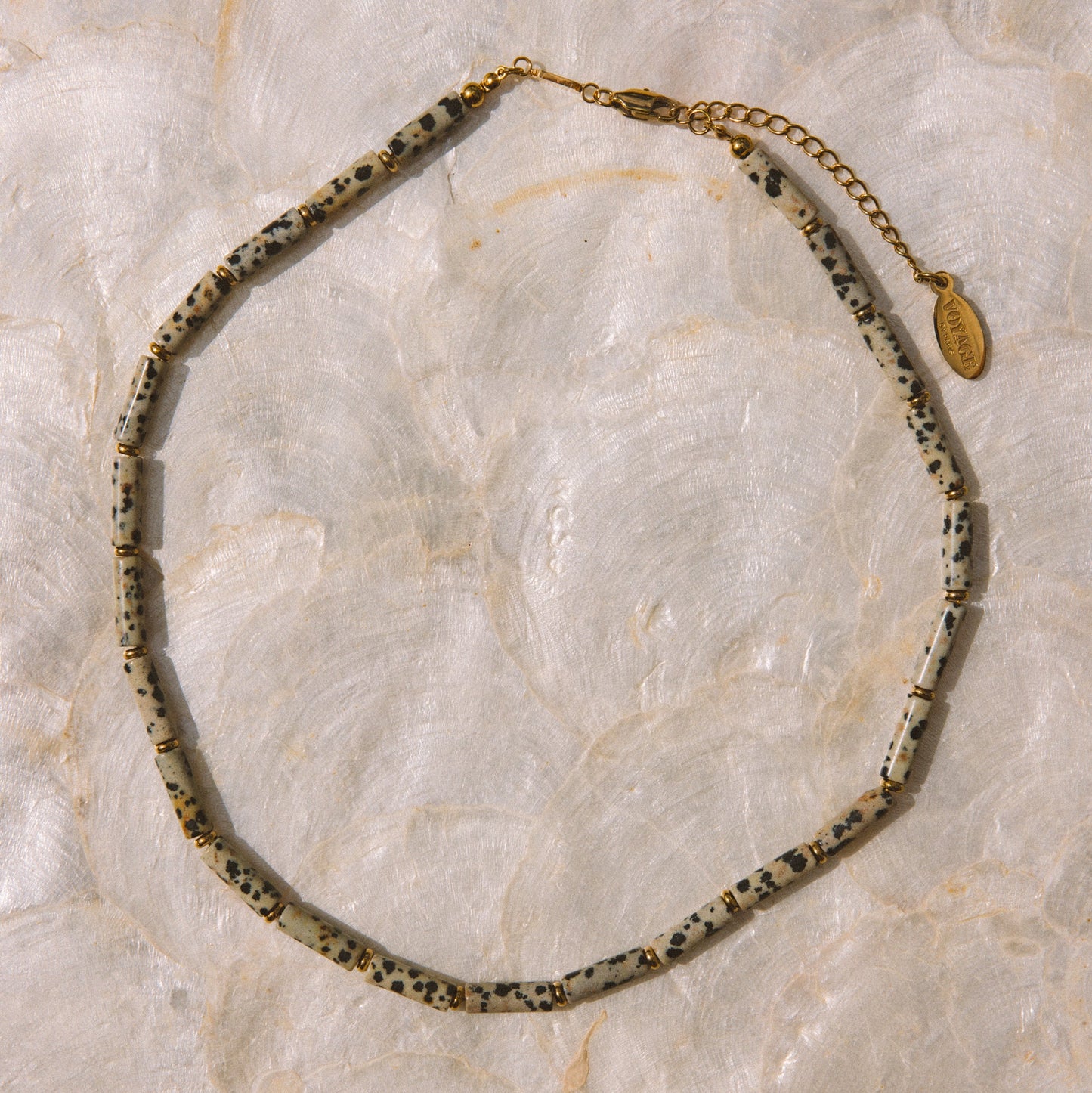 A beaded necklace with irregular speckled beads in black, white, and gray, accented by a gold chain extender and a small gold tag, displayed on a light beige marble surface.