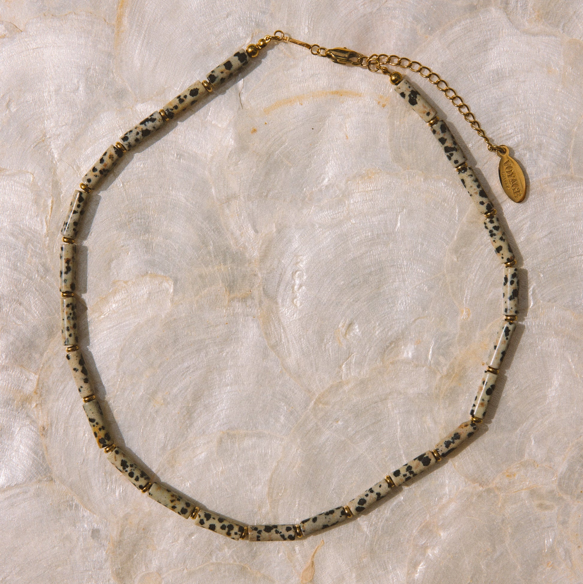 A beaded necklace with irregular speckled beads in black, white, and gray, accented by a gold chain extender and a small gold tag, displayed on a light beige marble surface.