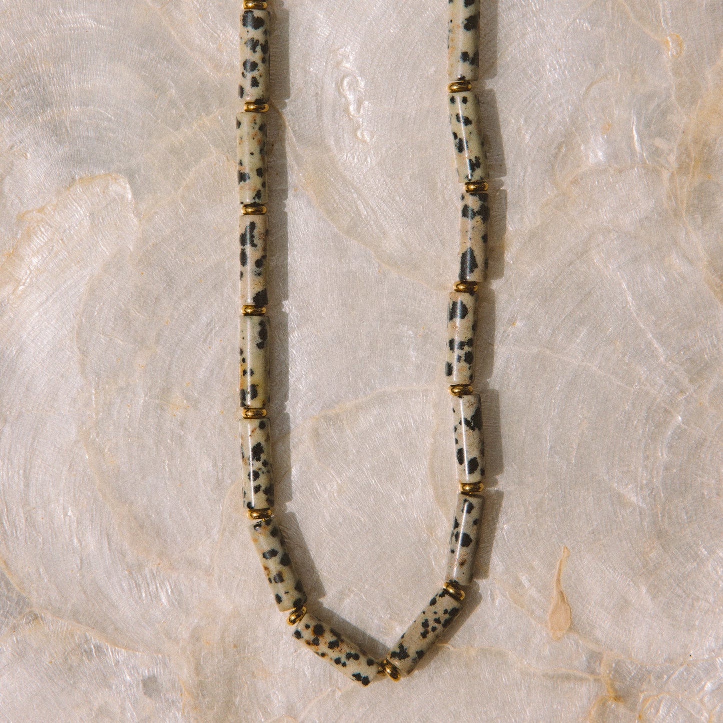 A beaded necklace made of elongated, rounded beads with a speckled black and white pattern on a cream base, separated by small gold spacer rings, laid flat on a light marble surface.