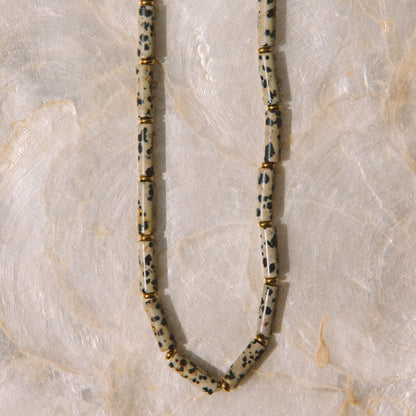 A beaded necklace made of elongated, rounded beads with a speckled black and white pattern on a cream base, separated by small gold spacer rings, laid flat on a light marble surface.