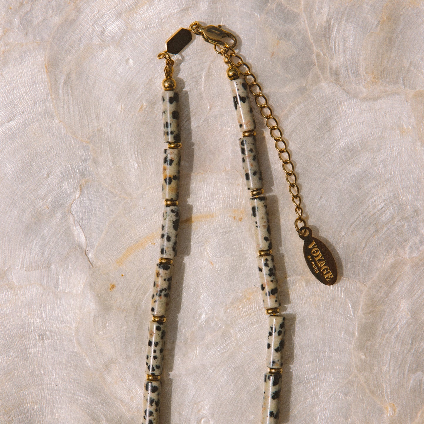 A beaded necklace featuring cream-colored cylindrical beads with black speckles, separated by small gold spacer rings, with a gold chain extender and a round gold tag engraved with 'Vogue 1980'.