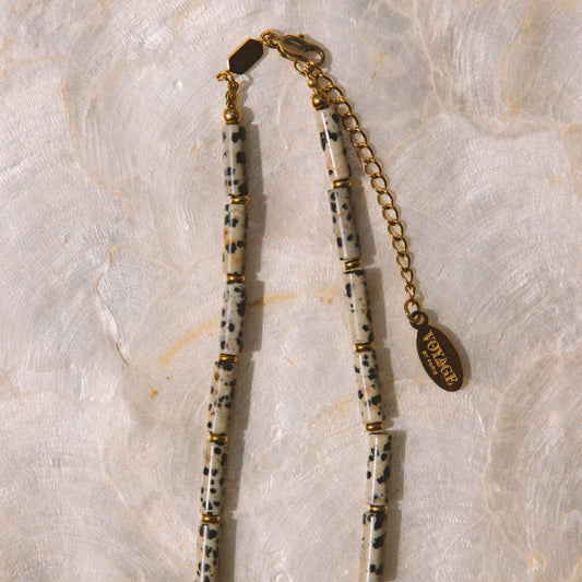 A beaded necklace featuring cream-colored cylindrical beads with black speckles, separated by small gold spacer rings, with a gold chain extender and a round gold tag engraved with 'Vogue 1980'.
