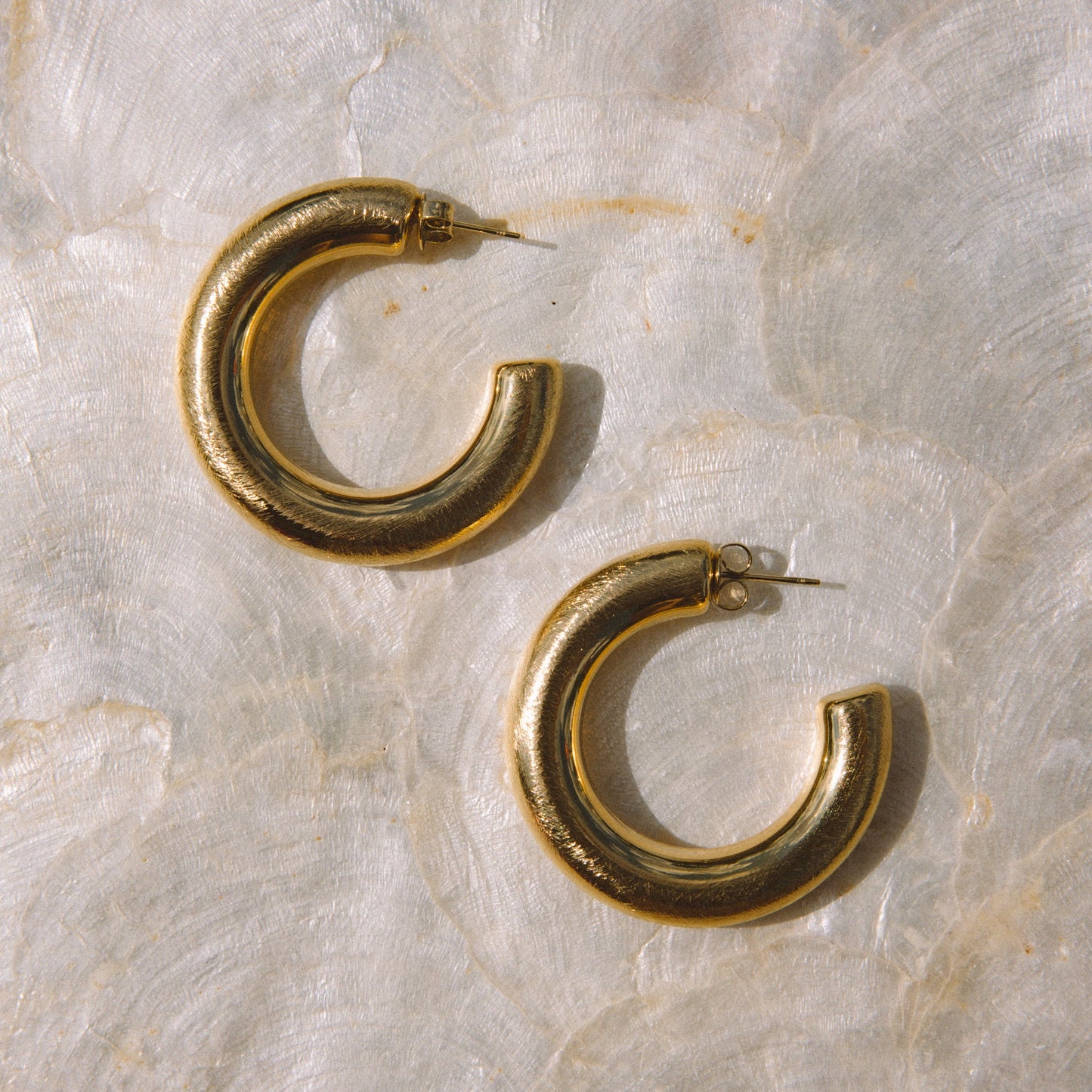 A pair of chunky, open-ended gold hoop earrings resting on a textured, light beige surface.
