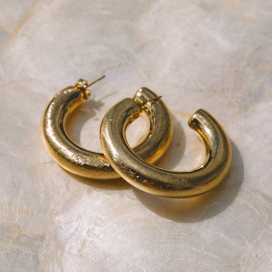 A pair of chunky, textured gold hoop earrings resting on a light beige marbled surface.