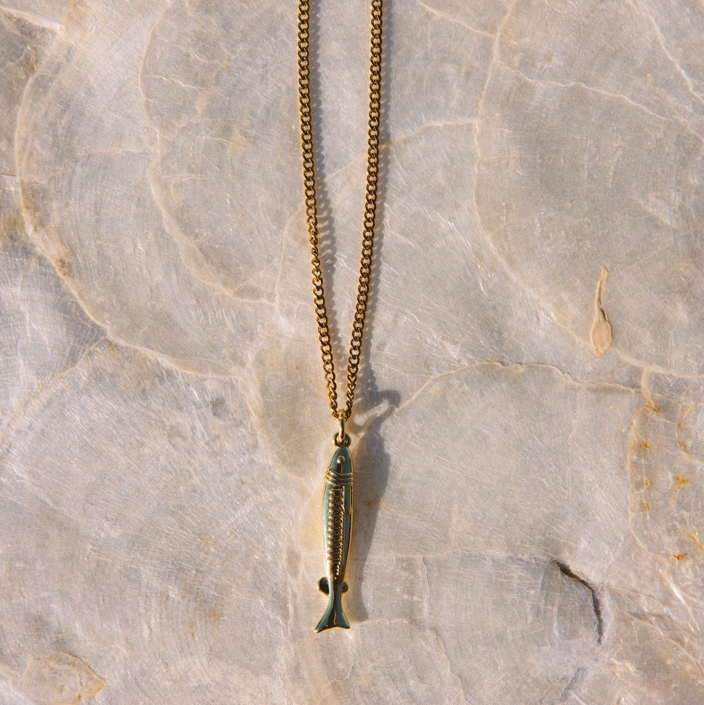 A gold-toned chain necklace with a small fish-shaped pendant, laid flat on a textured light beige stone surface.