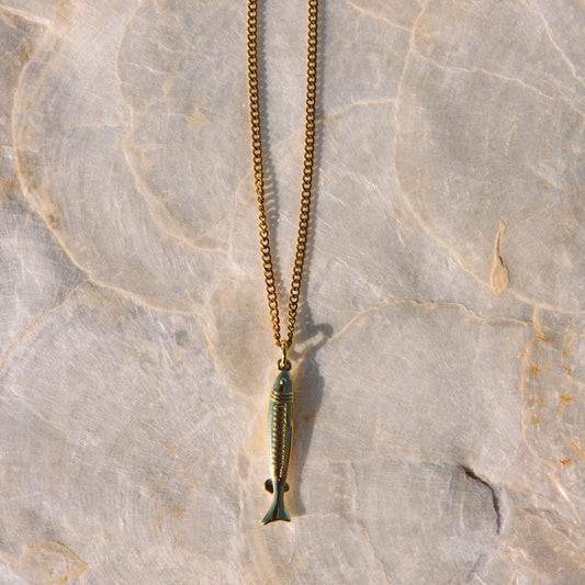 A gold-toned chain necklace with a small fish-shaped pendant, laid flat on a textured light beige stone surface.