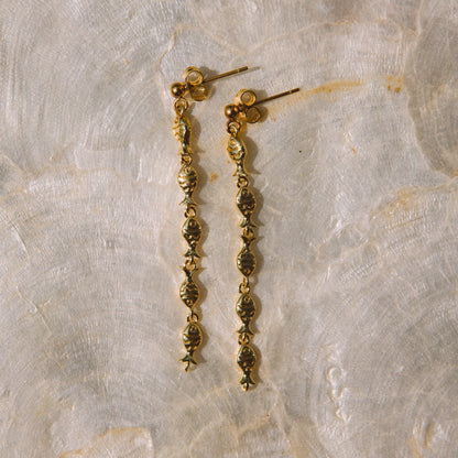 A pair of gold dangle earrings with textured, beaded drop chains resting on a textured beige stone surface.