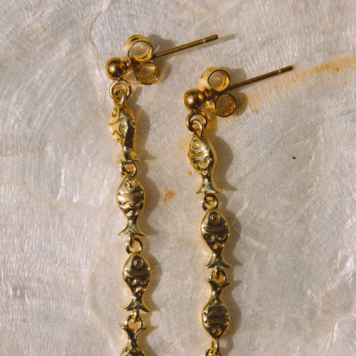 A pair of gold dangle earrings with textured, beaded drop chains resting on a textured beige stone surface.