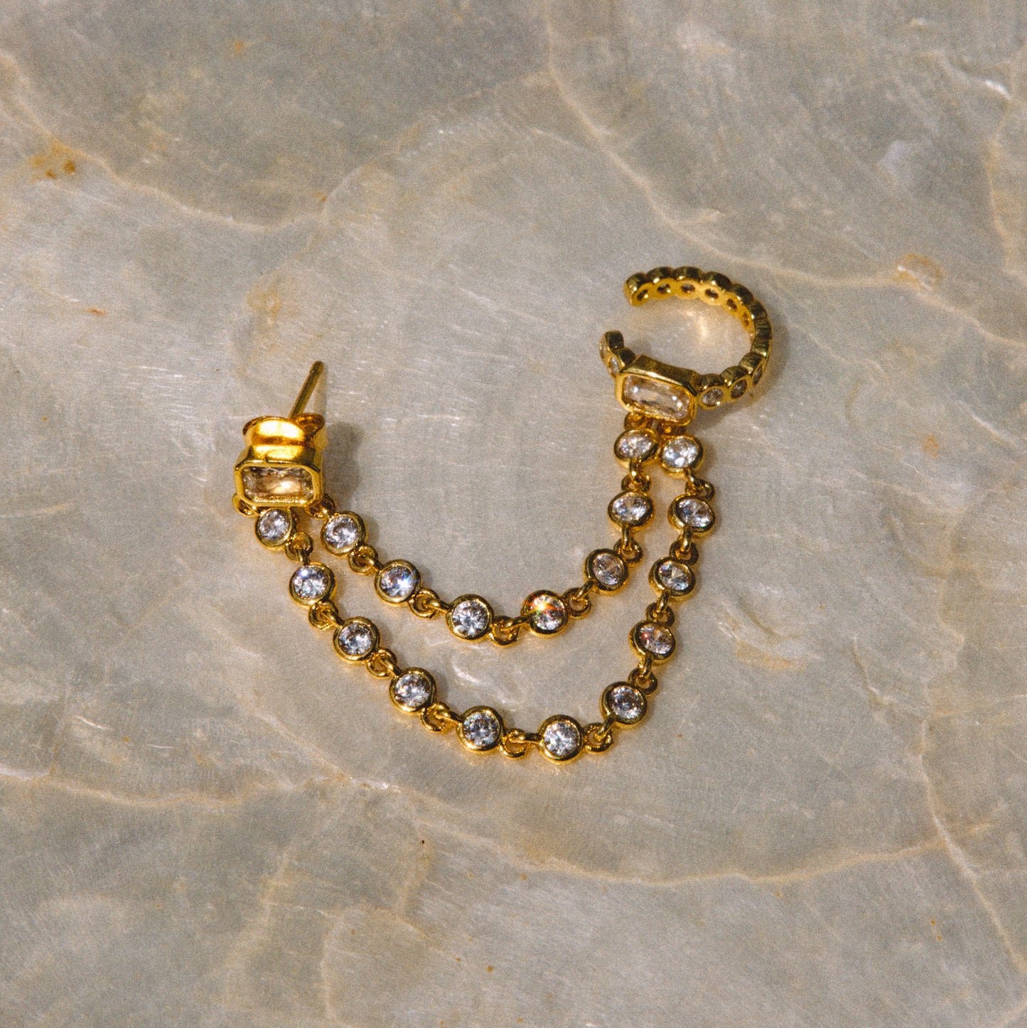 A gold huggie hoop earring with a dangling chain of clear round-cut rhinestones, displayed on a beige marble surface.