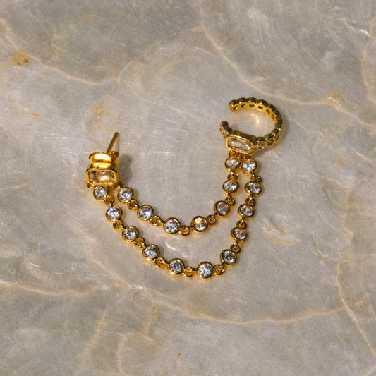 A gold huggie hoop earring with a dangling chain of clear round-cut rhinestones, displayed on a beige marble surface.