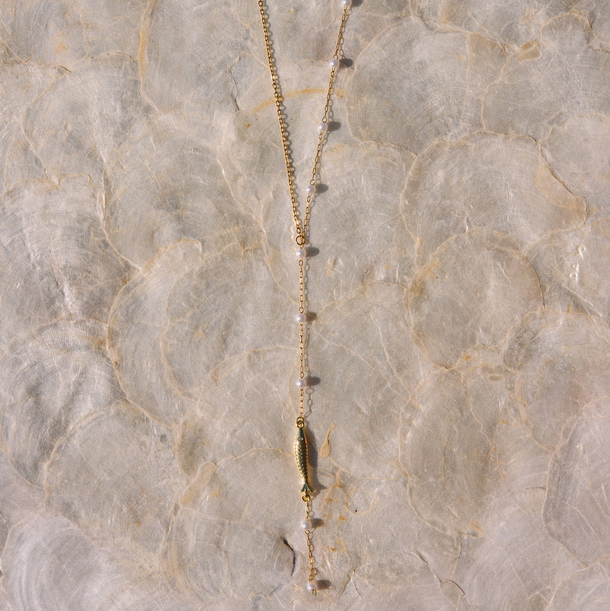A gold-toned lariat necklace with a central bar pendant and small bead accents, laid flat on a textured beige surface.