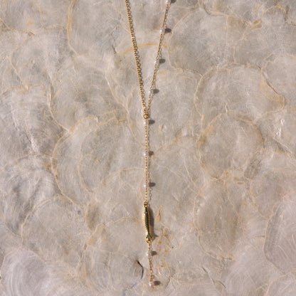 A gold-toned lariat necklace with a central bar pendant and small bead accents, laid flat on a textured beige surface.