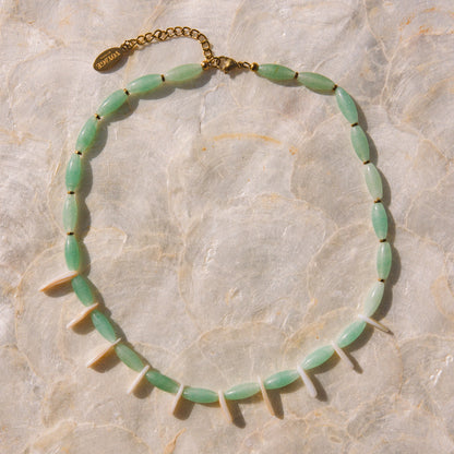 A beaded necklace with light green oval beads and white pointed accents, laid flat on a light beige stone-textured background. A gold-tone chain extender and clasp are visible at the top.