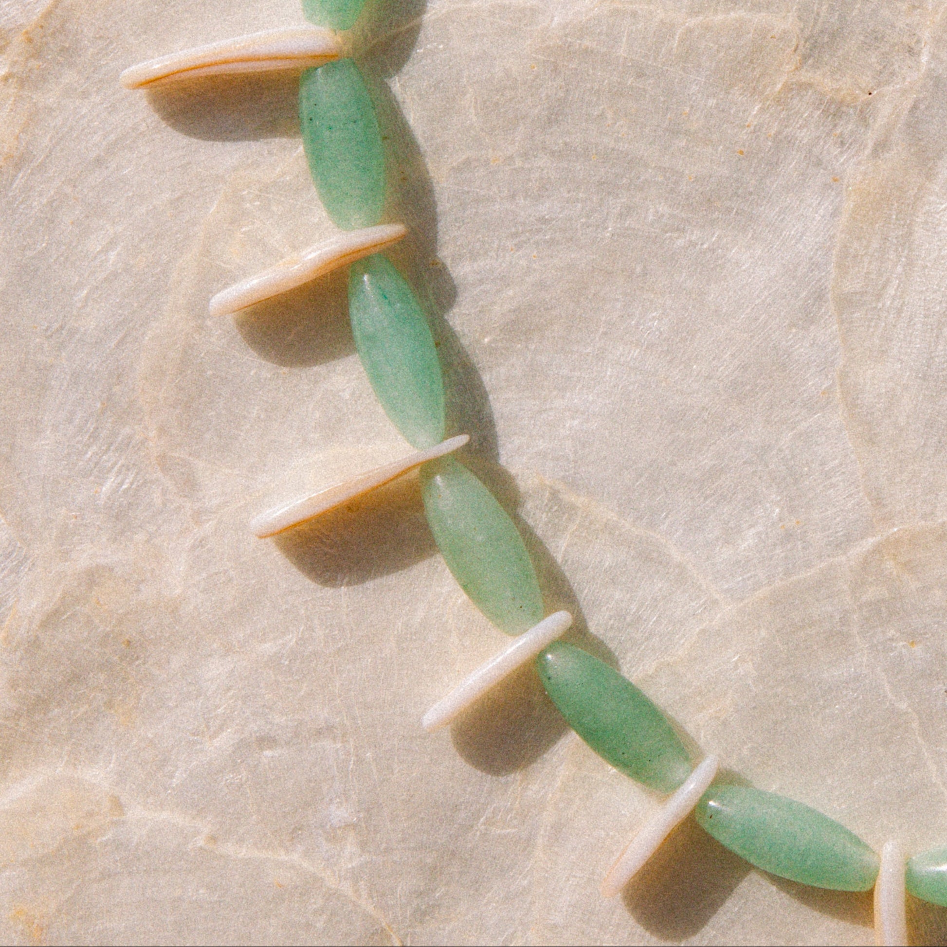A close-up of a beaded strand featuring elongated light green jade-like beads alternating with small cream-colored disc beads, laid diagonally on a textured beige surface.