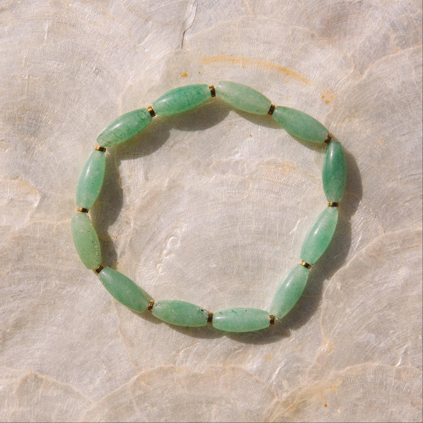 A beaded stretch bracelet made of polished light green jade-like oval beads separated by tiny dark spacers, displayed on a textured beige stone surface.