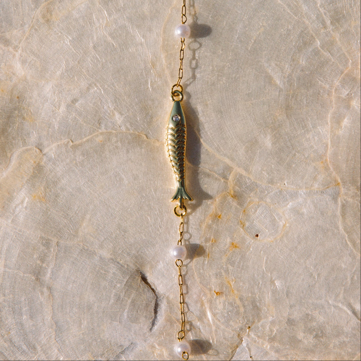 A delicate gold lariat necklace featuring a central gold fish charm, small white pearl beads, and a dangling gold bead, laid flat on a textured beige stone surface.