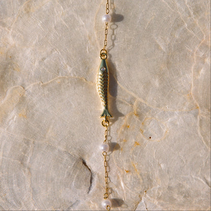 A delicate gold lariat necklace featuring a central gold fish charm, small white pearl beads, and a dangling gold bead, laid flat on a textured beige stone surface.