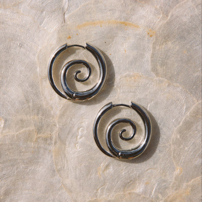 A pair of dark metallic spiral hoop earrings resting on a textured beige stone surface.