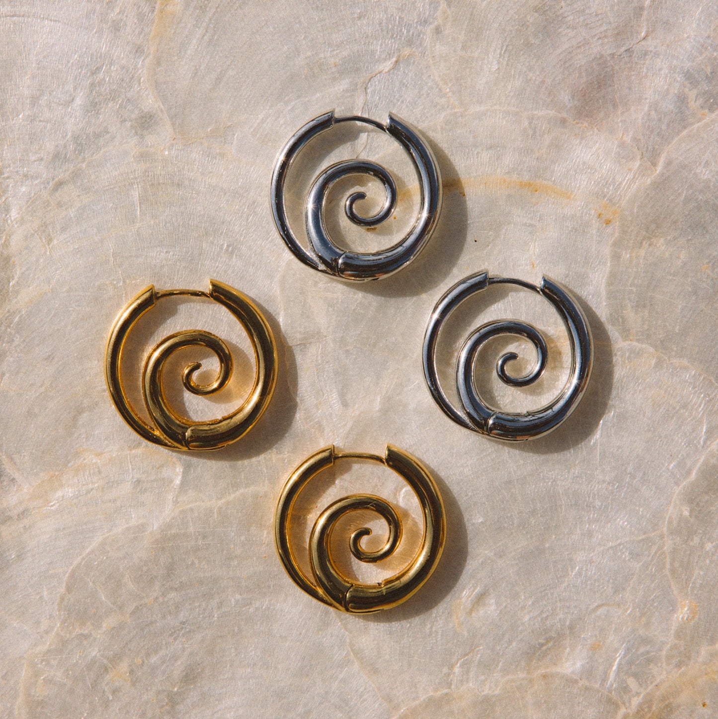 Four spiral-shaped hoop earrings in polished gold and dark silver tones, arranged on a light beige marble surface.