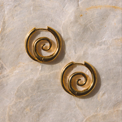A pair of gold spiral hoop earrings displayed on a textured beige stone surface.