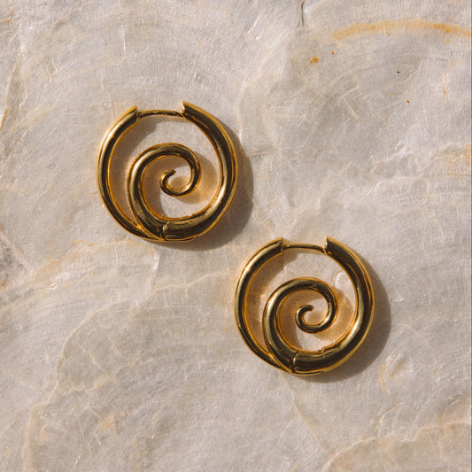 A pair of gold spiral hoop earrings displayed on a textured beige stone surface.