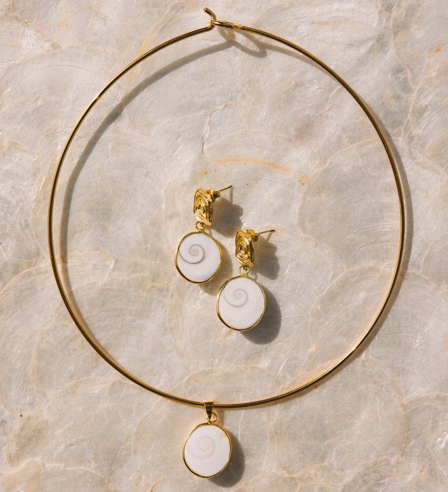 Gold jewelry set featuring a large hoop necklace and matching stud earrings with white spiral shell-like pendants, arranged on a light beige marble surface.