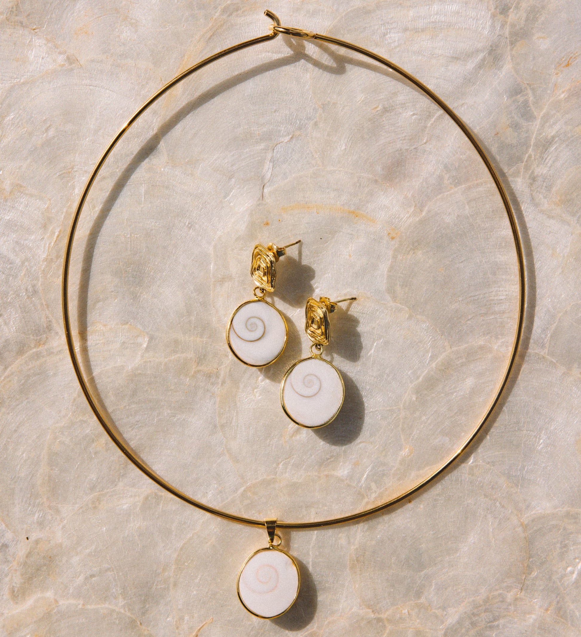 Gold jewelry set featuring a large hoop necklace and matching stud earrings with white spiral shell-like pendants, arranged on a light beige marble surface.