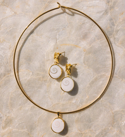 Gold jewelry set featuring a large hoop necklace and matching stud earrings with white spiral shell-like pendants, arranged on a light beige marble surface.