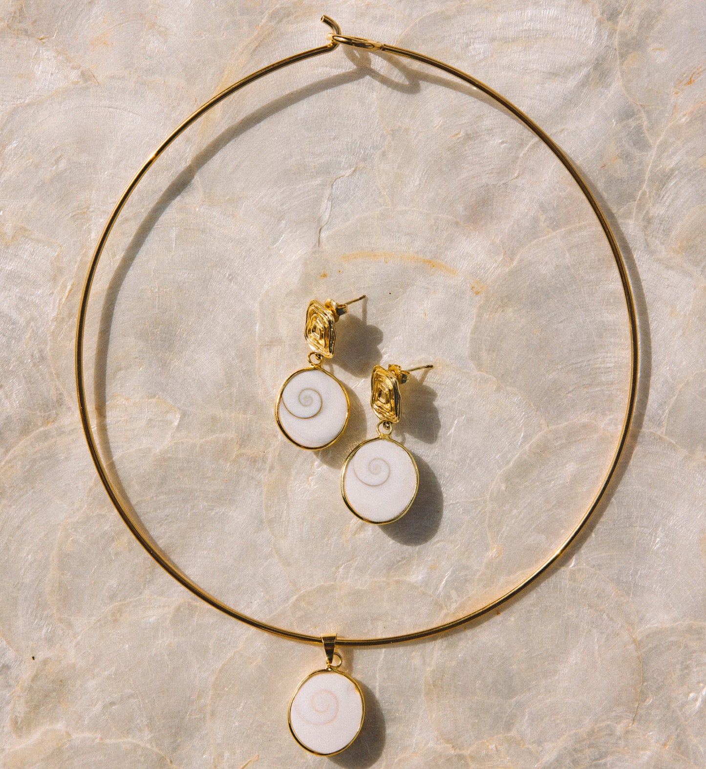 Gold jewelry set featuring a large hoop necklace and matching stud earrings with white spiral shell-like pendants, arranged on a light beige marble surface.