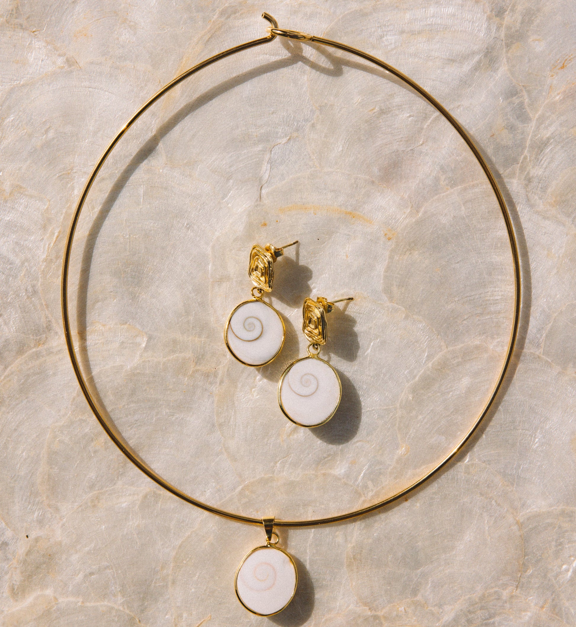 Gold jewelry set featuring a large hoop necklace and matching stud earrings with white spiral shell-like pendants, arranged on a light beige marble surface.
