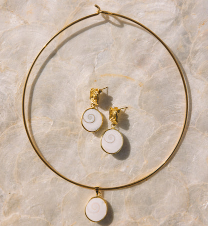 Gold jewelry set featuring a large hoop necklace and matching stud earrings with white spiral shell-like pendants, arranged on a light beige marble surface.