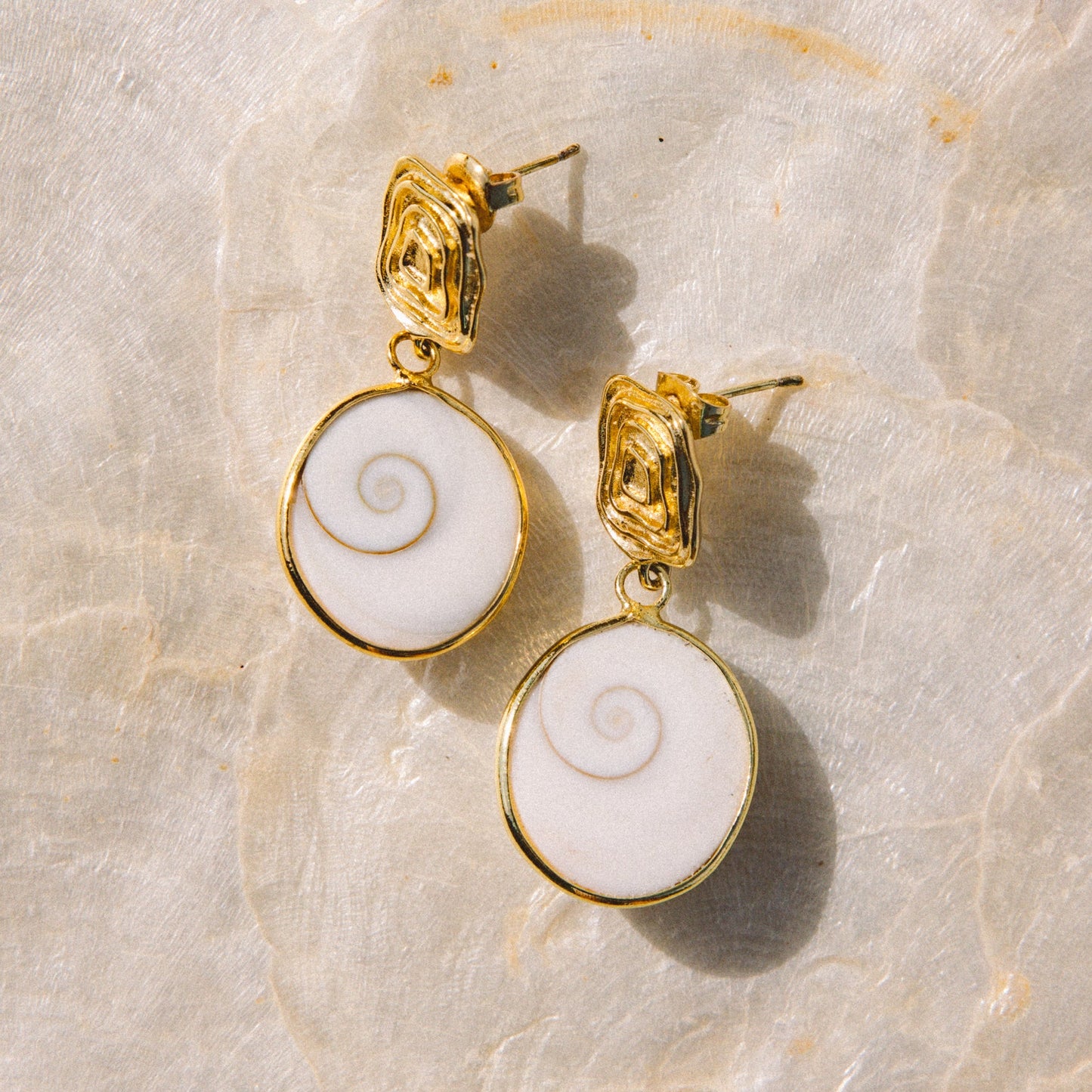 A pair of gold drop earrings with oval white inlay featuring a spiral design, displayed on a light beige stone surface.