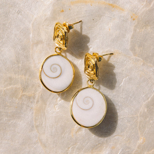 A pair of gold drop earrings with oval white inlay featuring a spiral design, displayed on a light beige stone surface.